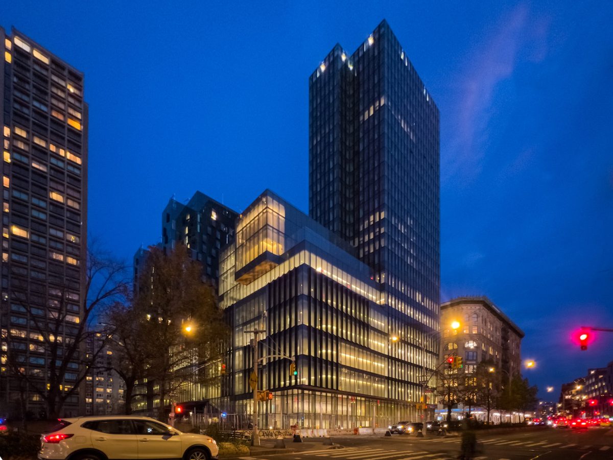 A street view at night of a tall geometric building with lights shining through the windows.