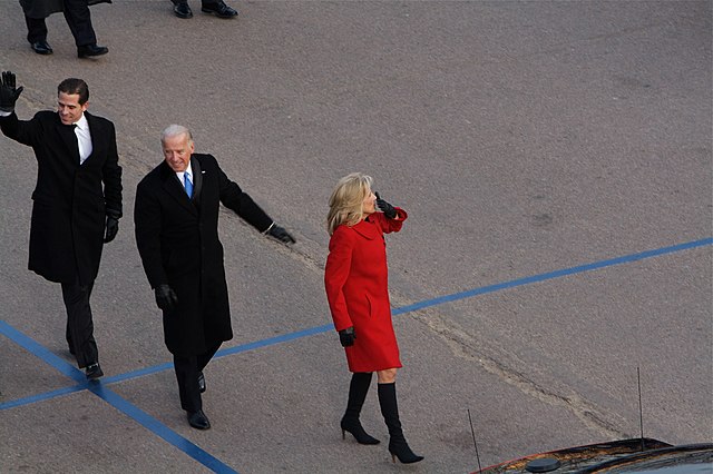 First lady Jill Biden, wearing a red dress and black boots, holds a cellphone as President Joe Biden, wearing a black suit with a blue tie, and his son Hunter Biden, wearing a black shirt, walk behind her.