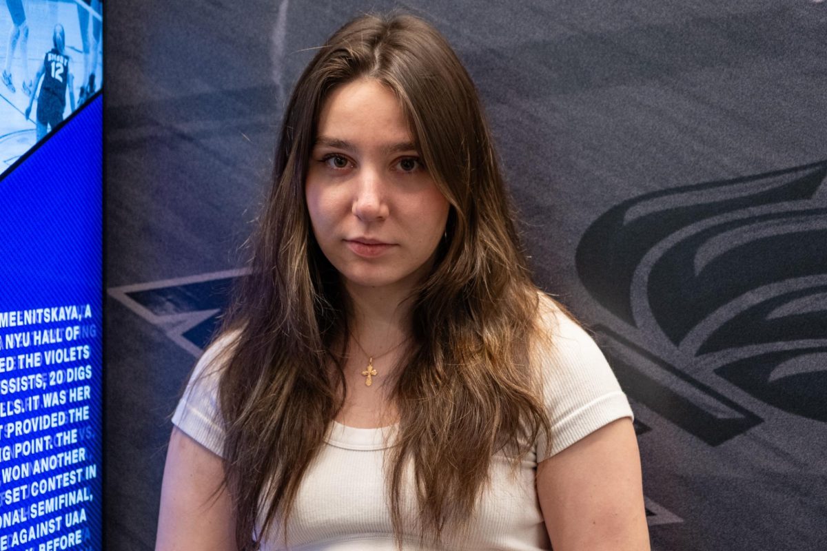 A woman in a white shirt stands in front of a dark background.