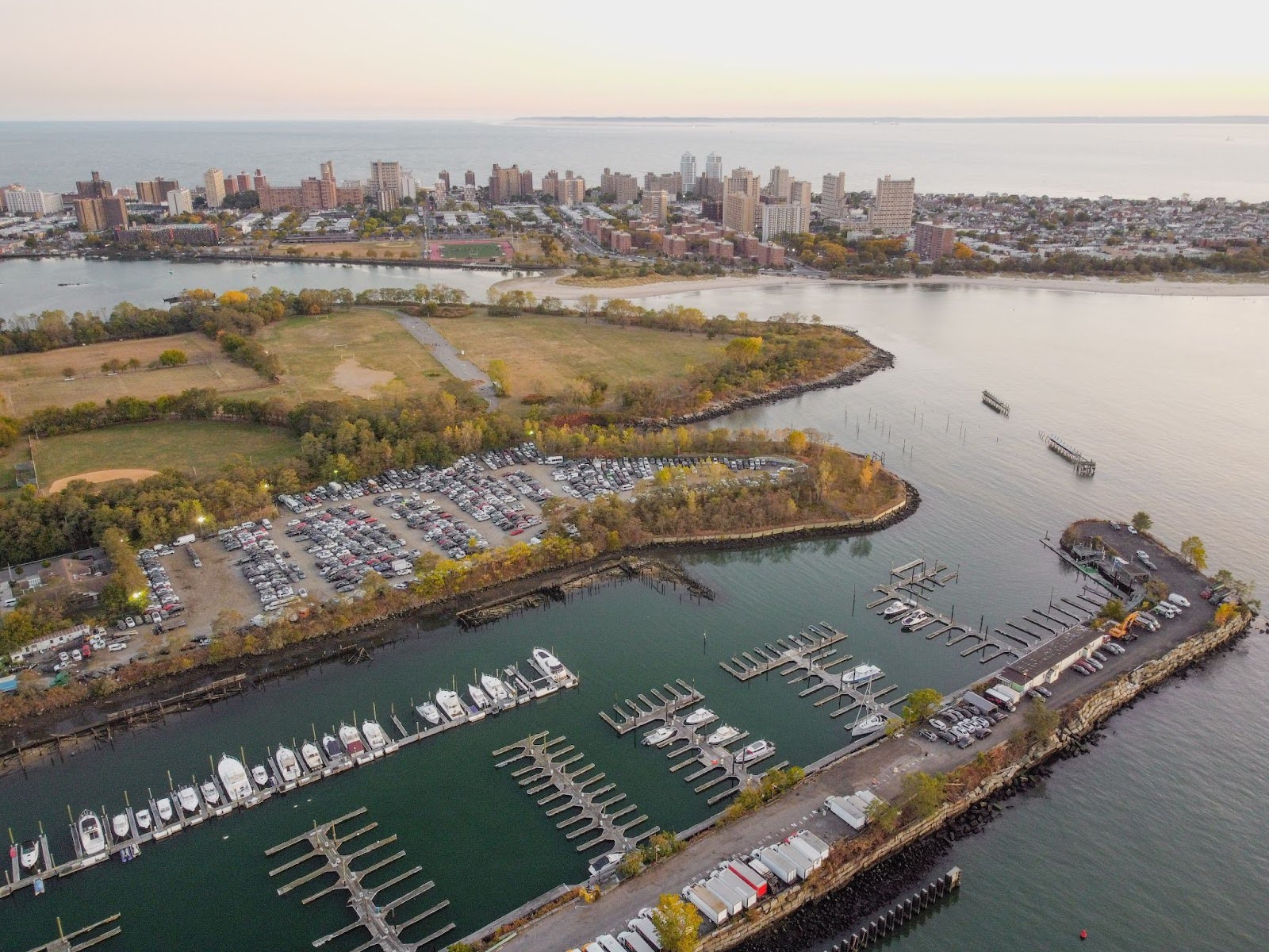 An aerial view of a harbor with ships and parking lots next to it and a city skyline in the background.