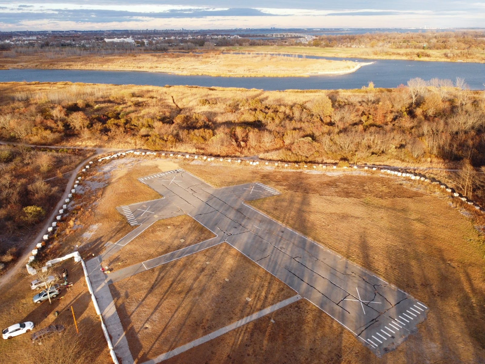 An aerial view of an old airfield with a cracked runway, surrounded by autumn trees and a calm body of water in the background.