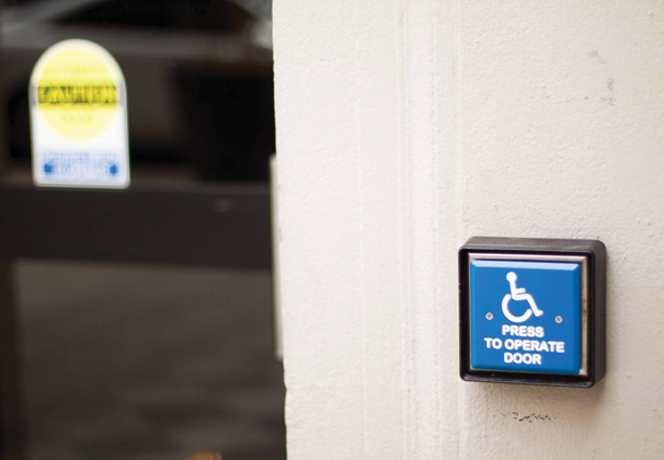A photo of a doorway on campus with a blue accessibility button that reads Press to Operate Door to the right.