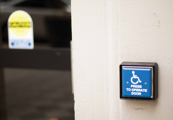 A photo of a doorway on campus with a blue accessibility button that reads "Press to Operate Door" to the right.